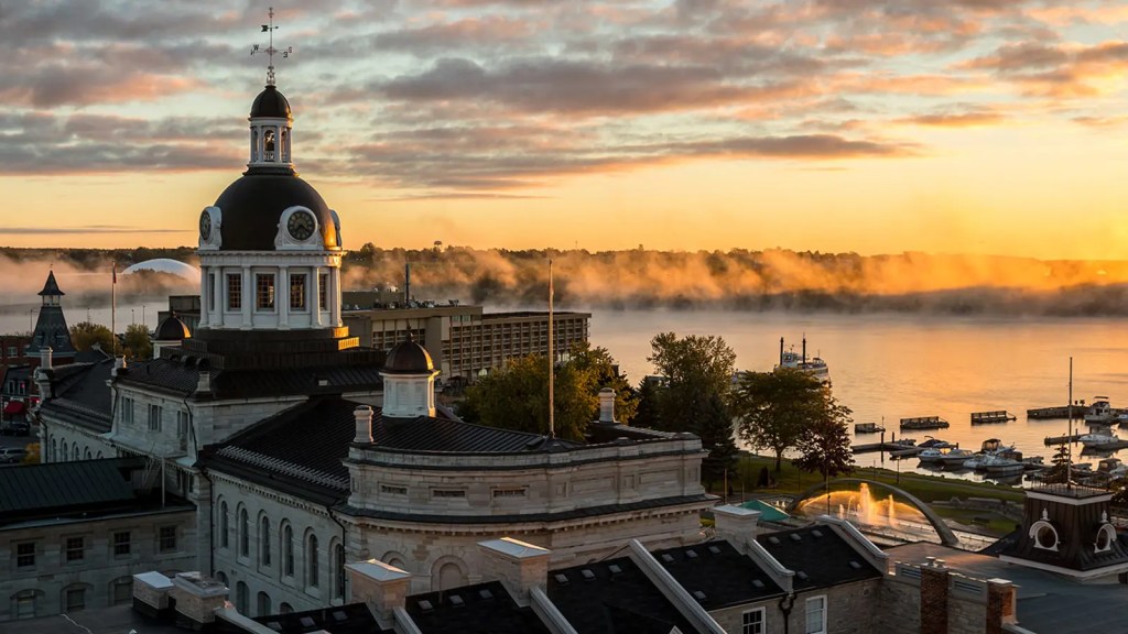 A scenic view of the waterfront in Kingston, Ontario, featuring historic buildings with a prominent dome at sunrise, surrounded by mist on the water.
